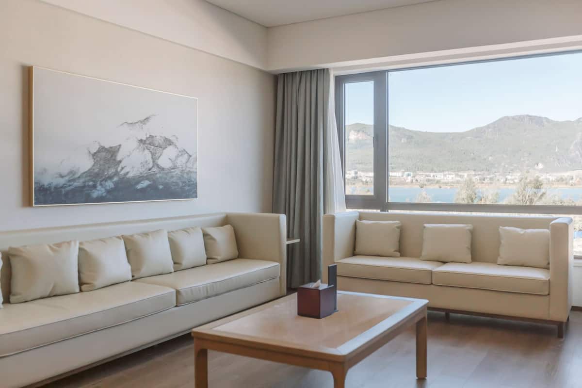 A modern living room with two beige sofas, a wooden coffee table, and a large window offering a view of a lake and mountains.