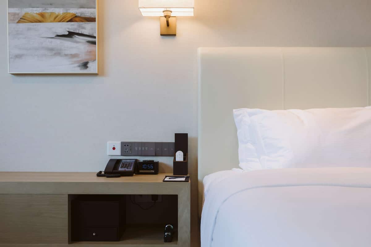 A hotel room with a bed, a bedside table with a phone, a lamp, and a painting on the wall.