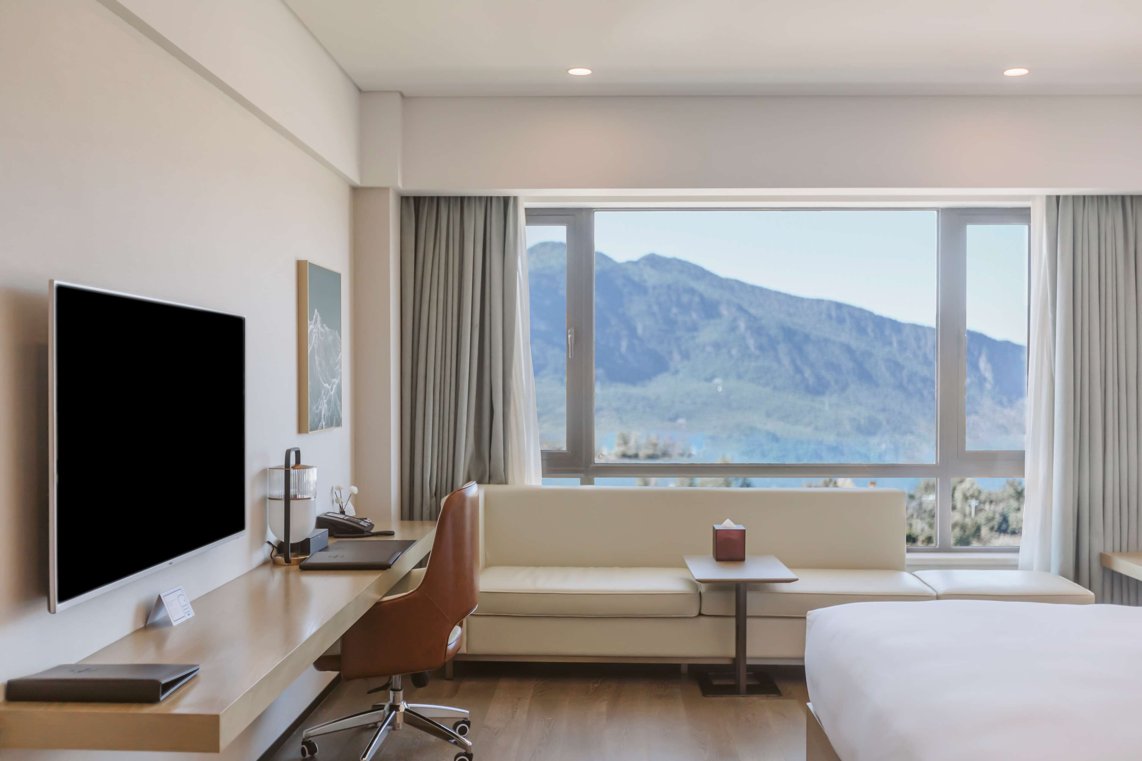 A hotel room with a bed, a desk with a flat-screen TV, a sofa, and a window offering a mountain view.