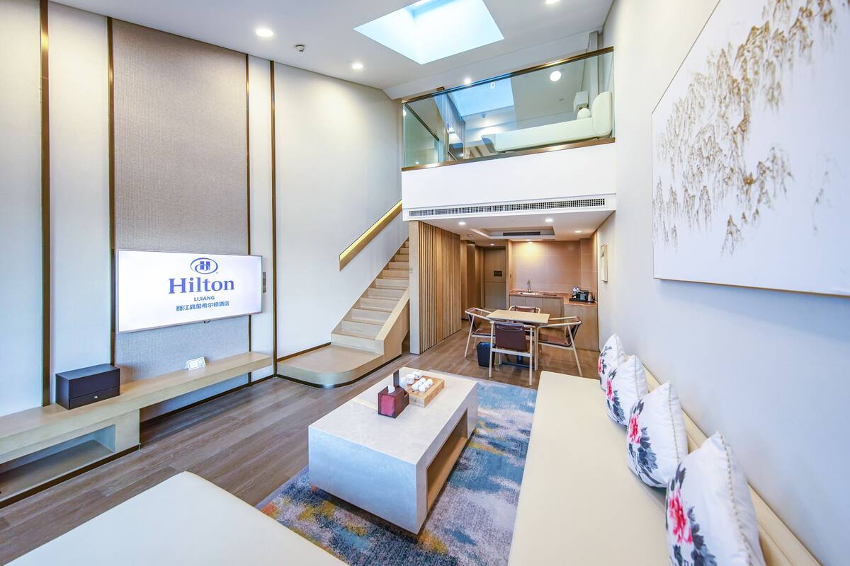 A modern hotel lobby with a high ceiling, a staircase, a seating area, and a dining space.