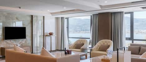Habitación de hotel moderna con un televisor de pantalla plana grande, asientos cómodos, una mesita de café y vistas a un lago y montañas.