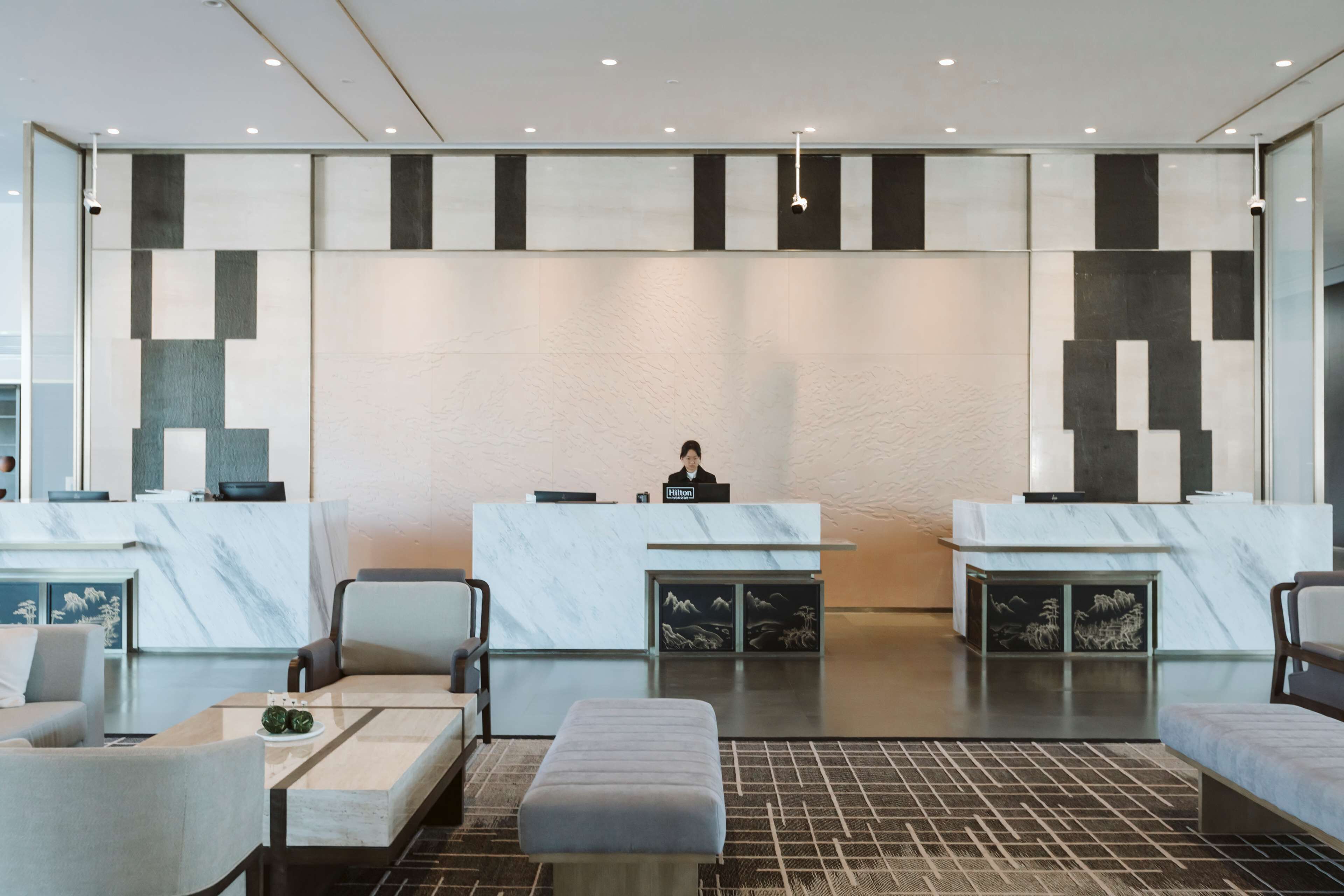 A modern hotel lobby with marble reception desks, seating areas, and a receptionist.