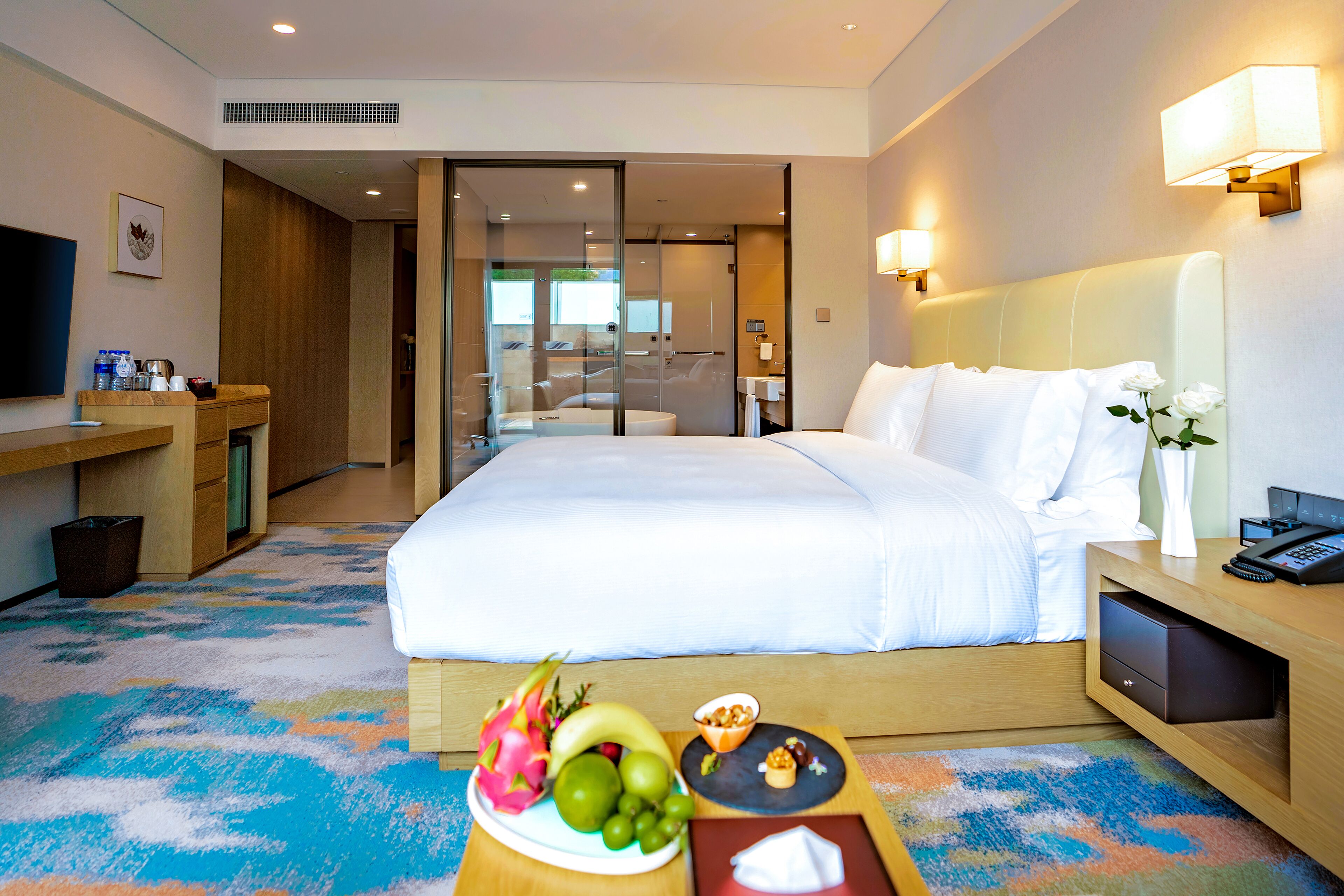 Hotelkamer met een groot bed, een bureau met een flatscreen-tv, een schaal met fruit en snacks, en een badkamer zichtbaar door een glazen deur.