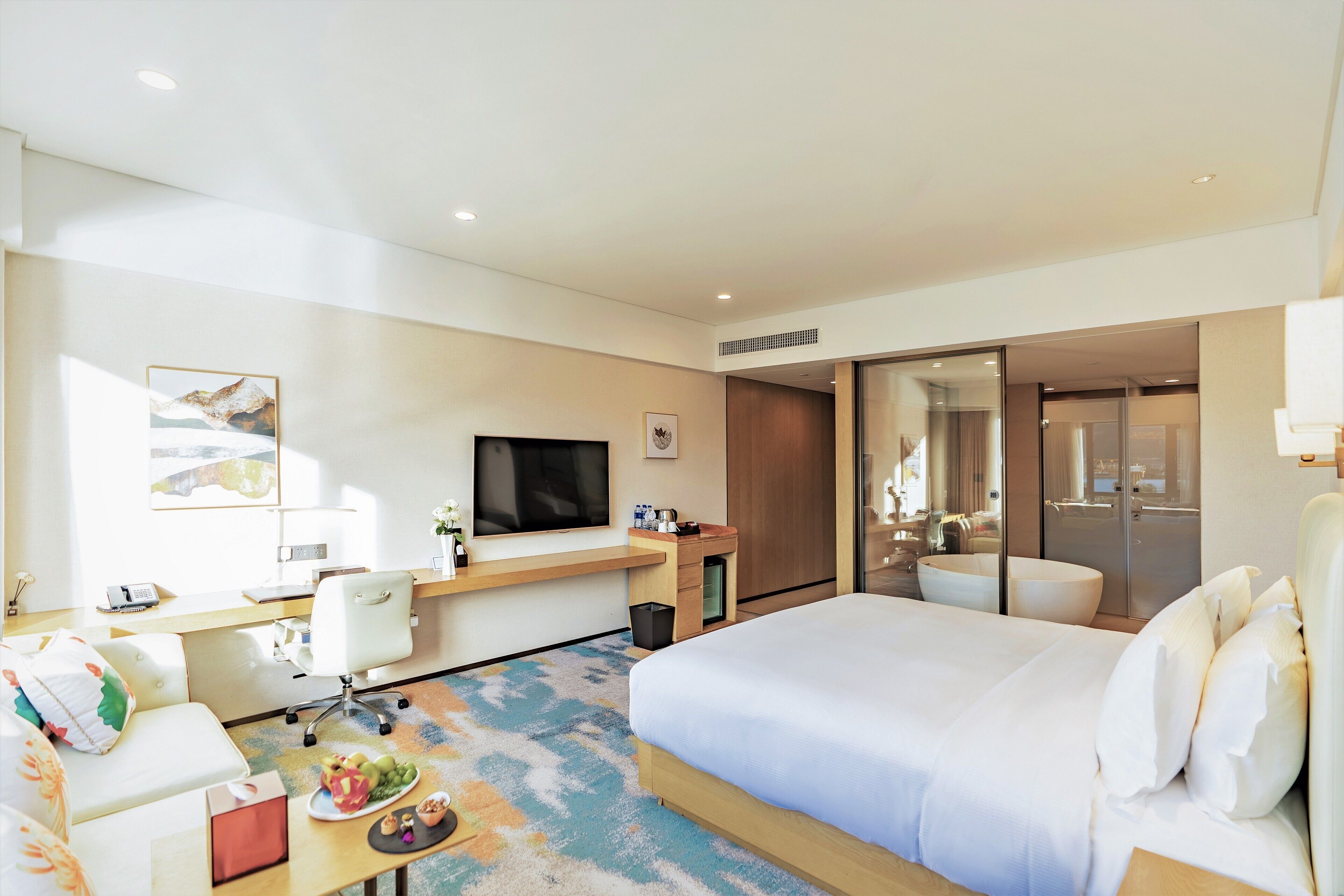 A modern hotel room with a large bed, a desk with a computer, a TV, and a seating area with a sofa and armchair.