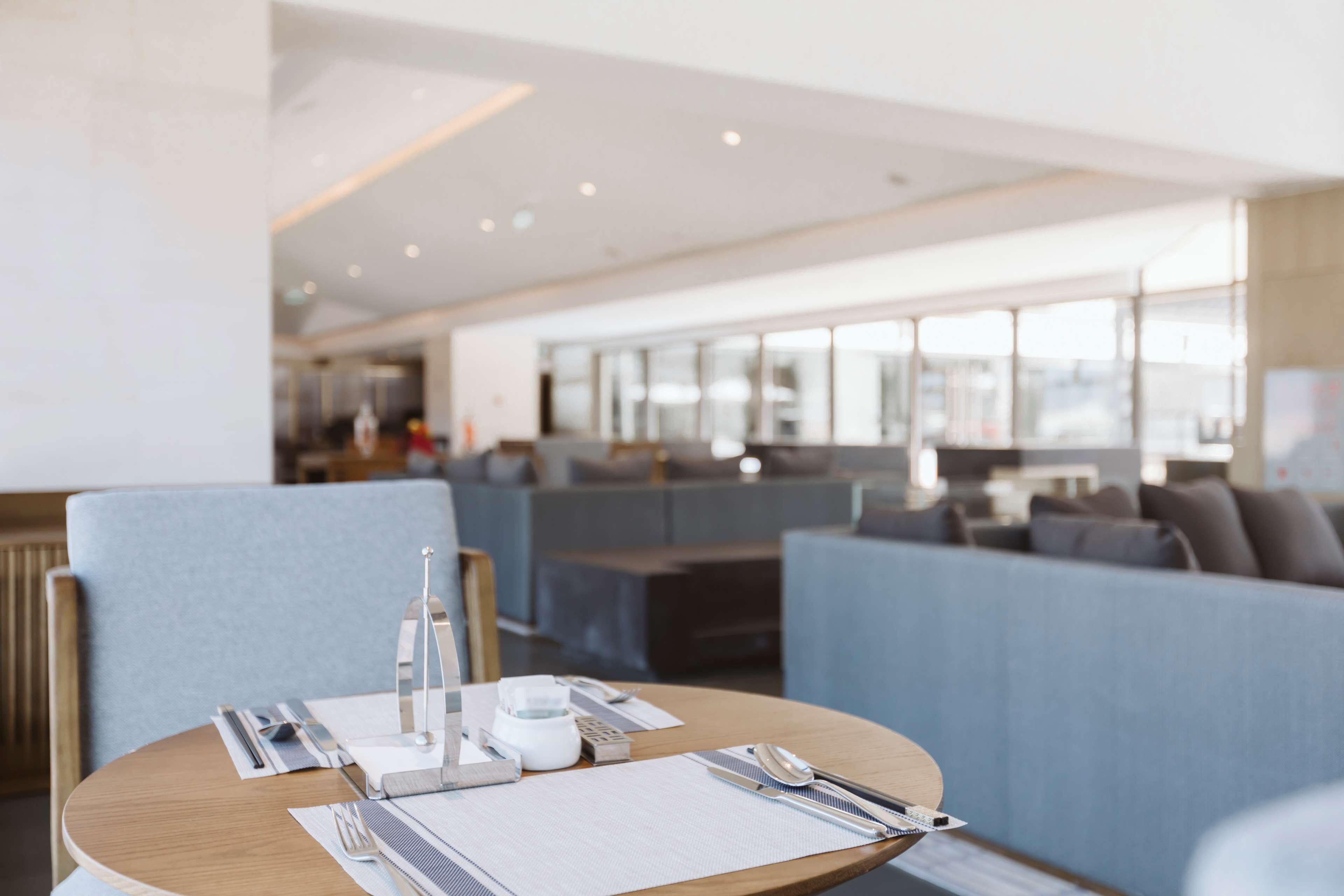 Un comedor moderno con una mesa redonda dispuesta para dos personas, que incluye un menú, cubiertos y una copa. La habitación tiene un diseño minimalista con asientos tapizados en gris y ventanas amplias que proporcionan luz natural.