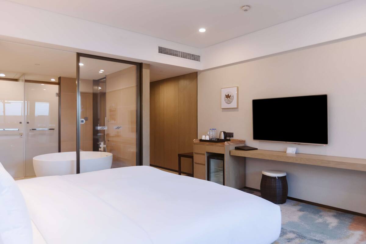 A modern hotel room with a large bed, a desk with a flat-screen TV, a small refrigerator, and a glass shower enclosure.