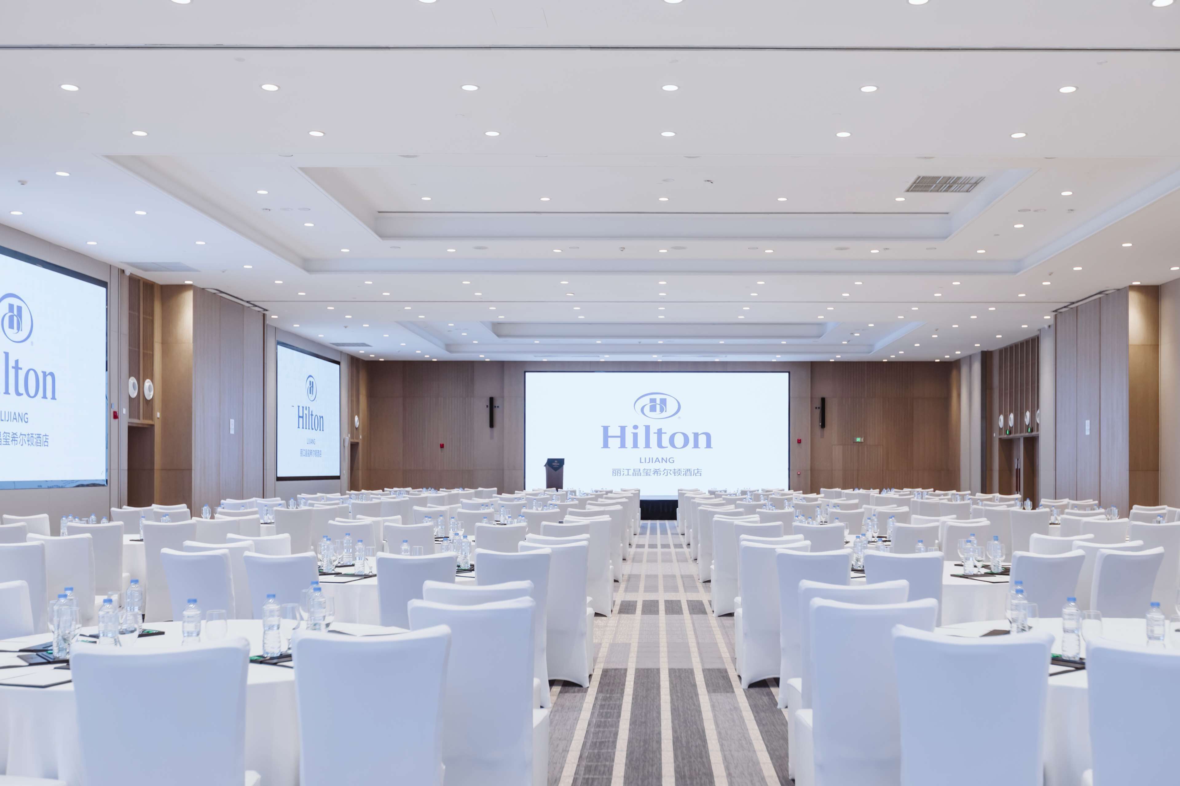 A conference room with rows of white tables and chairs, a large screen displaying the Hilton logo, and a podium at the front.