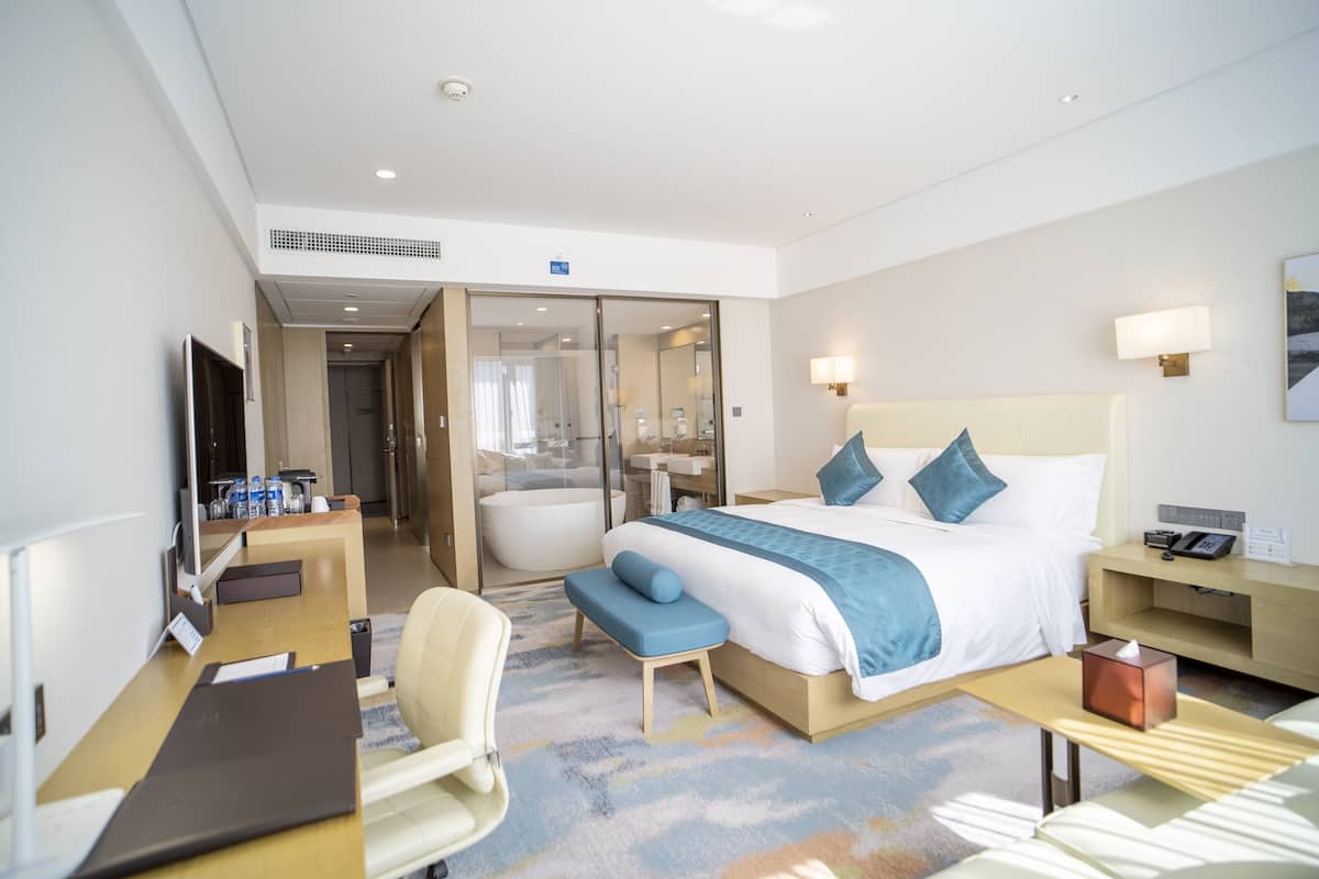 A hotel room with a bed, a desk, a chair, a bedside table, a lamp, and a bathroom visible through a glass door.