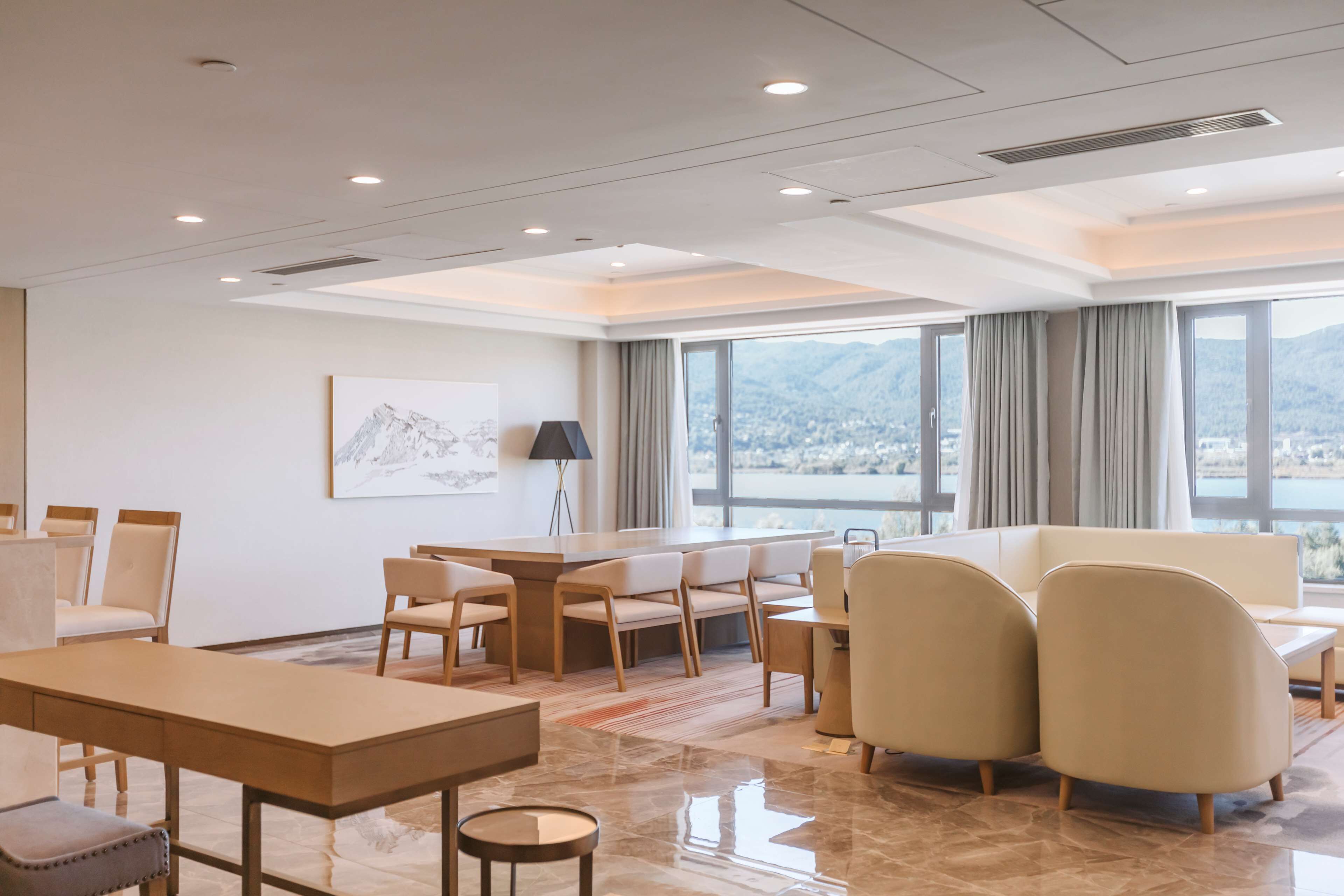 A modern hotel room with a large window offering a view of mountains and a cityscape. There is a dining area with a table and chairs, and a seating area with a sofa and armchairs.