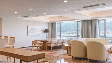 A modern hotel room with a large window offering a view of mountains and a cityscape. There is a dining area with a table and chairs, and a seating area with a sofa and armchairs.
