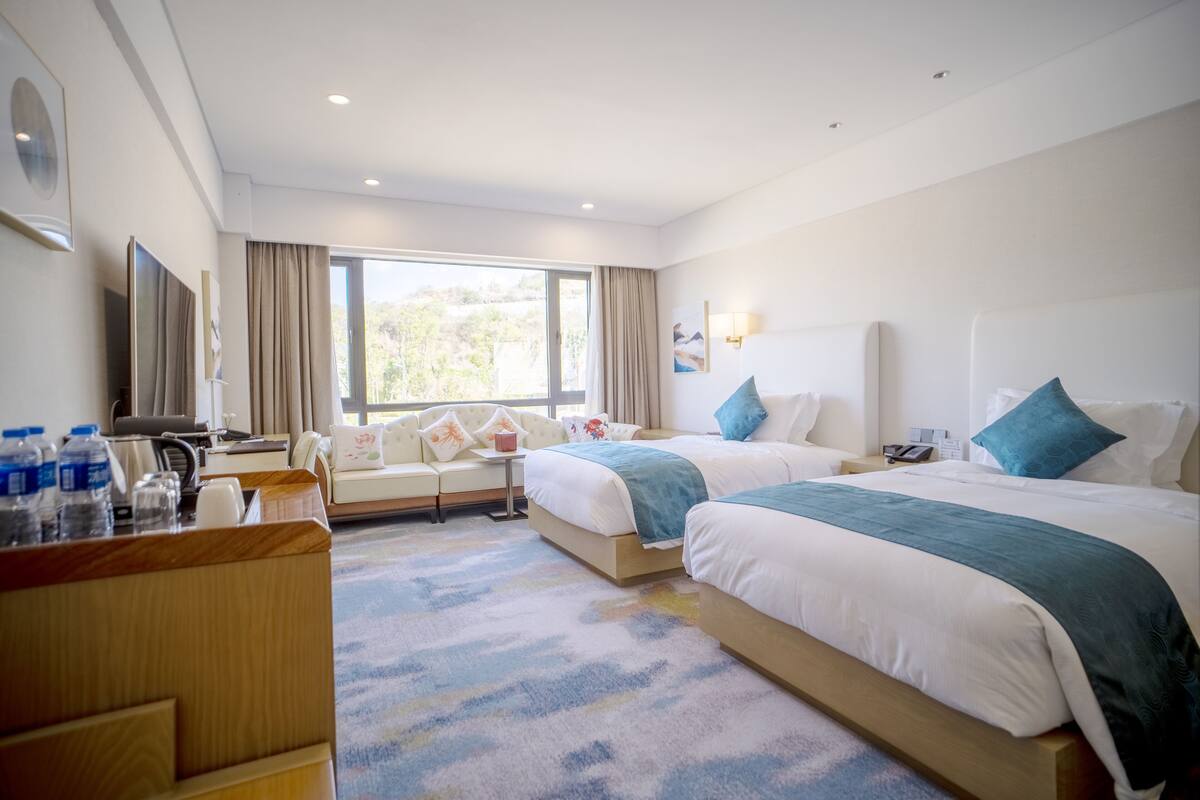 A hotel room with two beds, a desk, a TV, and a window with curtains.