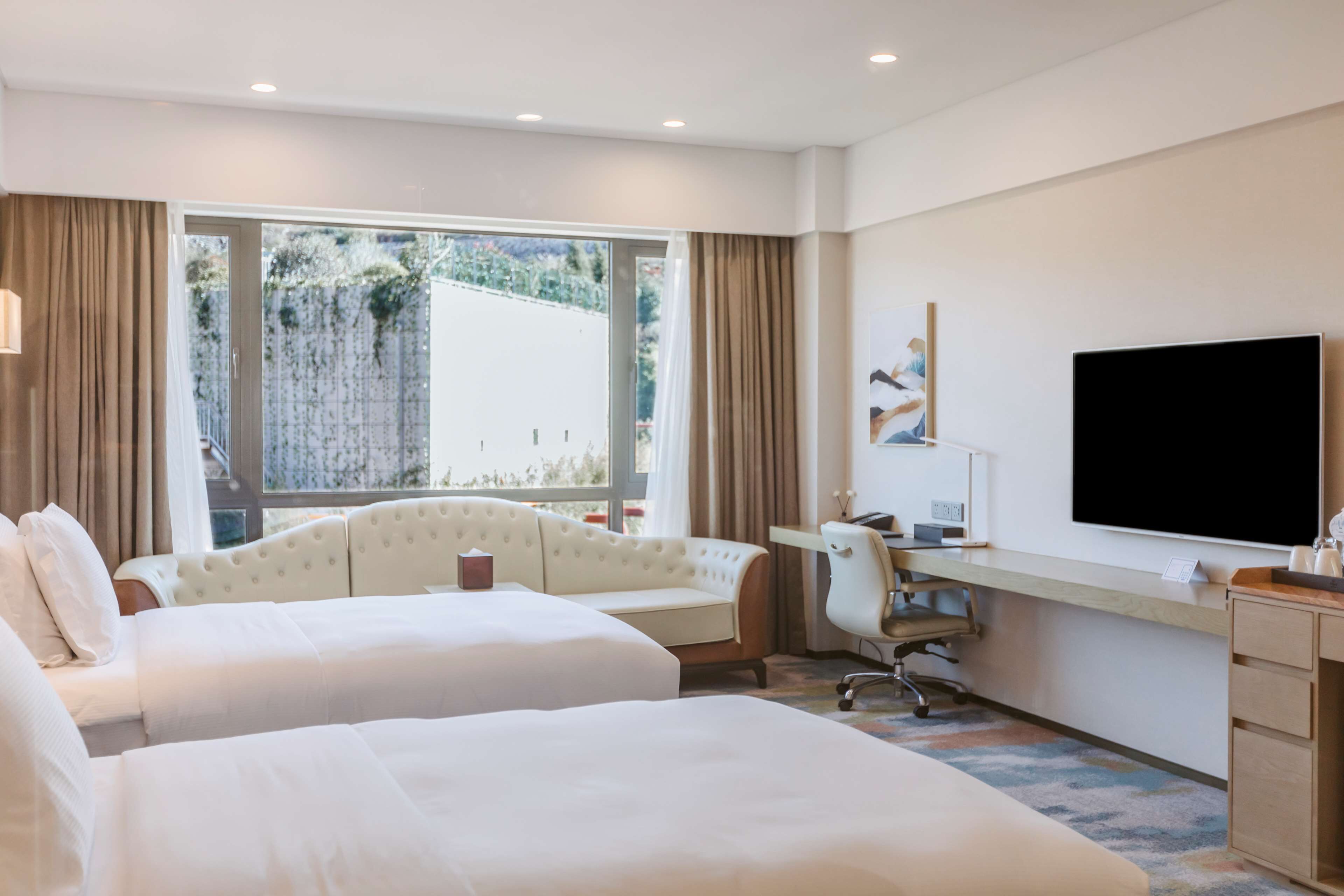 A hotel room with two beds, a desk, a chair, a flat-screen TV, and a large window offering a view of greenery.