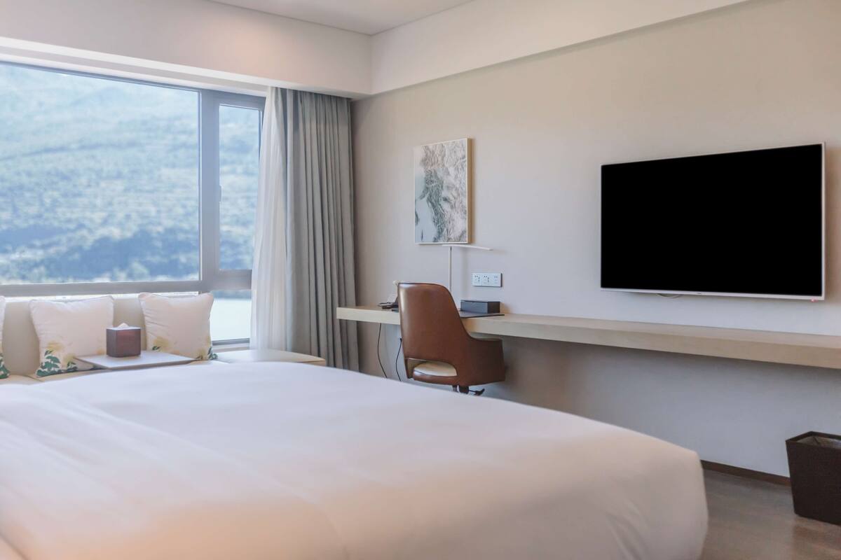 A hotel room with a large bed, a desk with a chair, a flat-screen TV, and a window with a view of the city.