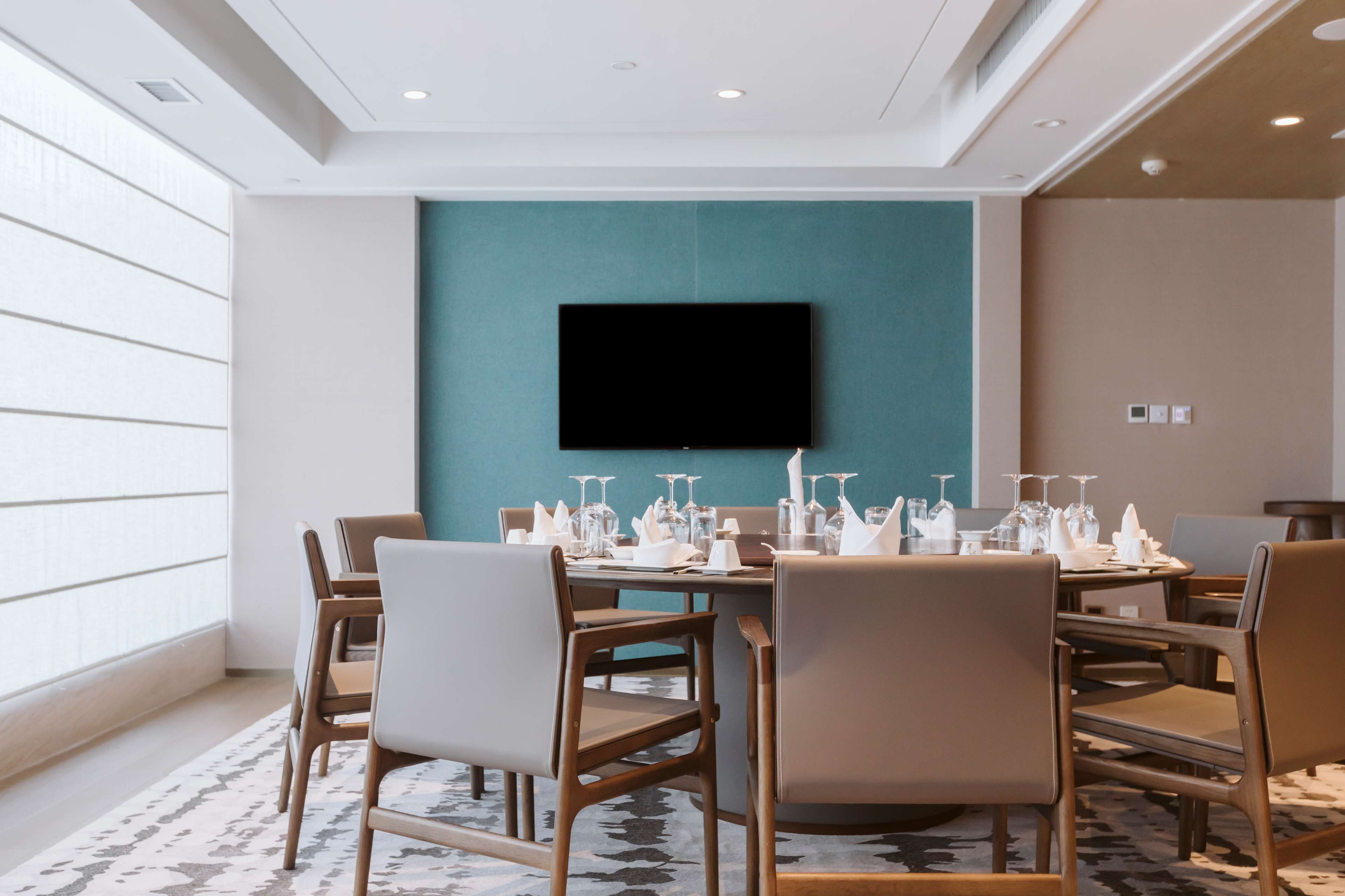 A hotel dining room with a teal accent wall, a flat-screen TV mounted above a table set for six with glasses and cutlery.