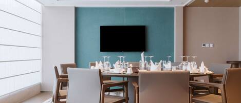 Un comedor de hotel con una pared de acento color turquesa, un televisor de pantalla plana montado sobre una mesa preparada para seis personas, con vasos y cubiertos.