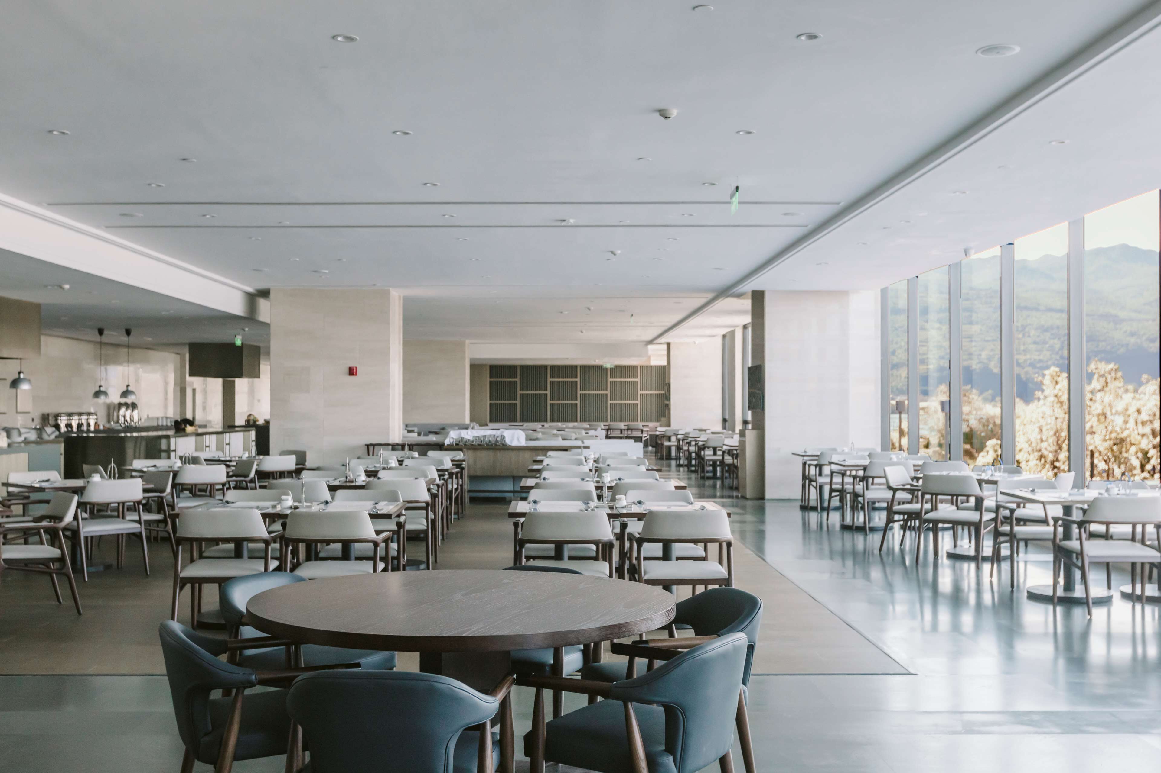 A spacious dining area with multiple tables and chairs, a kitchen in the background, and large windows offering a view of the outdoors.