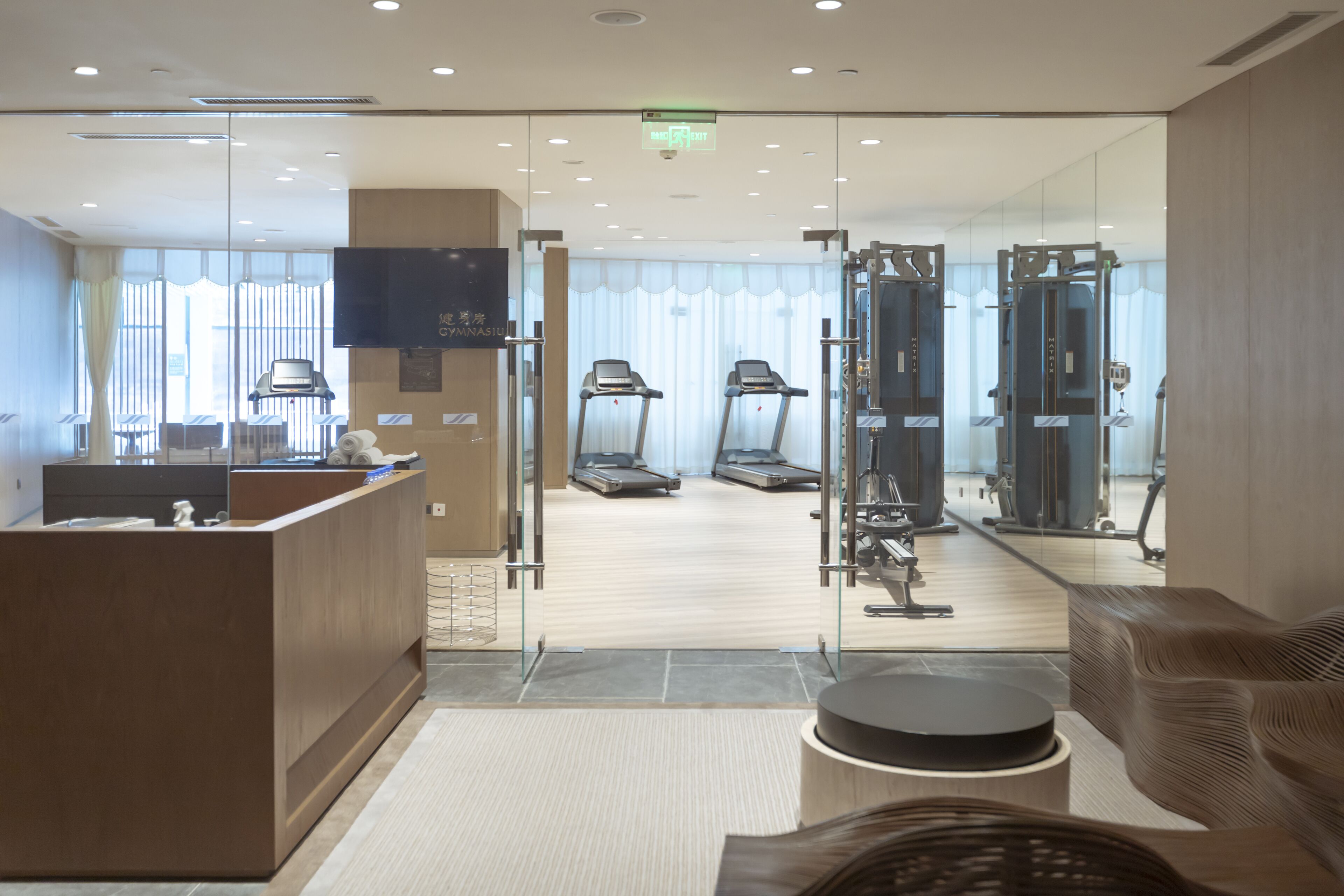 A modern hotel lobby with a reception desk, seating area, and a view of the gym area through glass doors.
