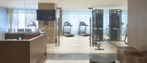 A modern hotel lobby with a reception desk, seating area, and a view of the gym area through glass doors.
