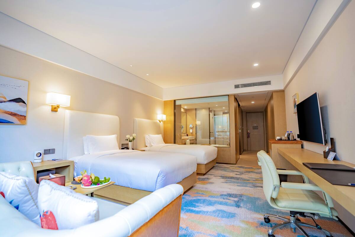 A hotel room with two beds, a desk, a chair, a TV, and a bathroom visible through a glass door.