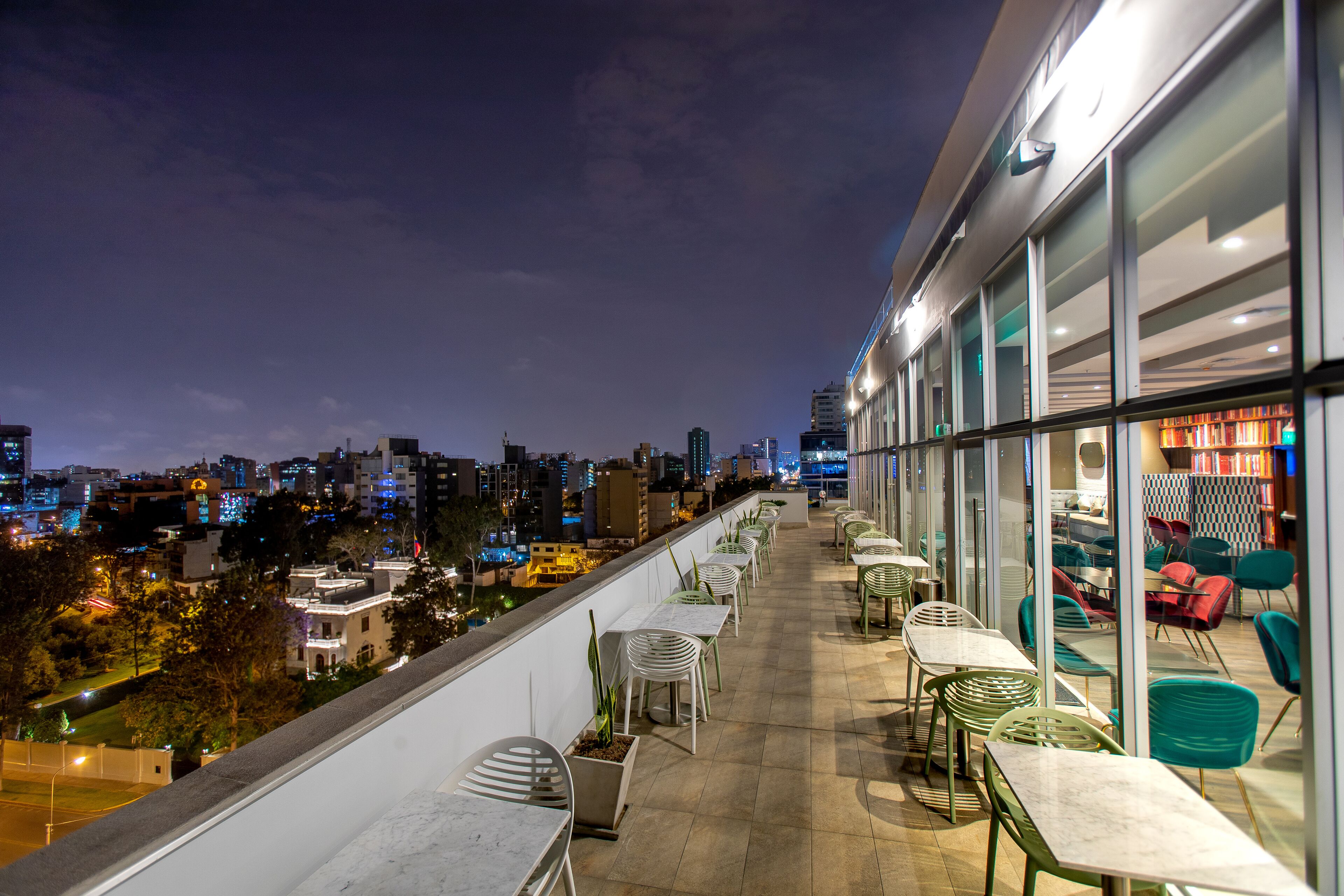 A rooftop terrace with tables and chairs, overlooking a cityscape at night.