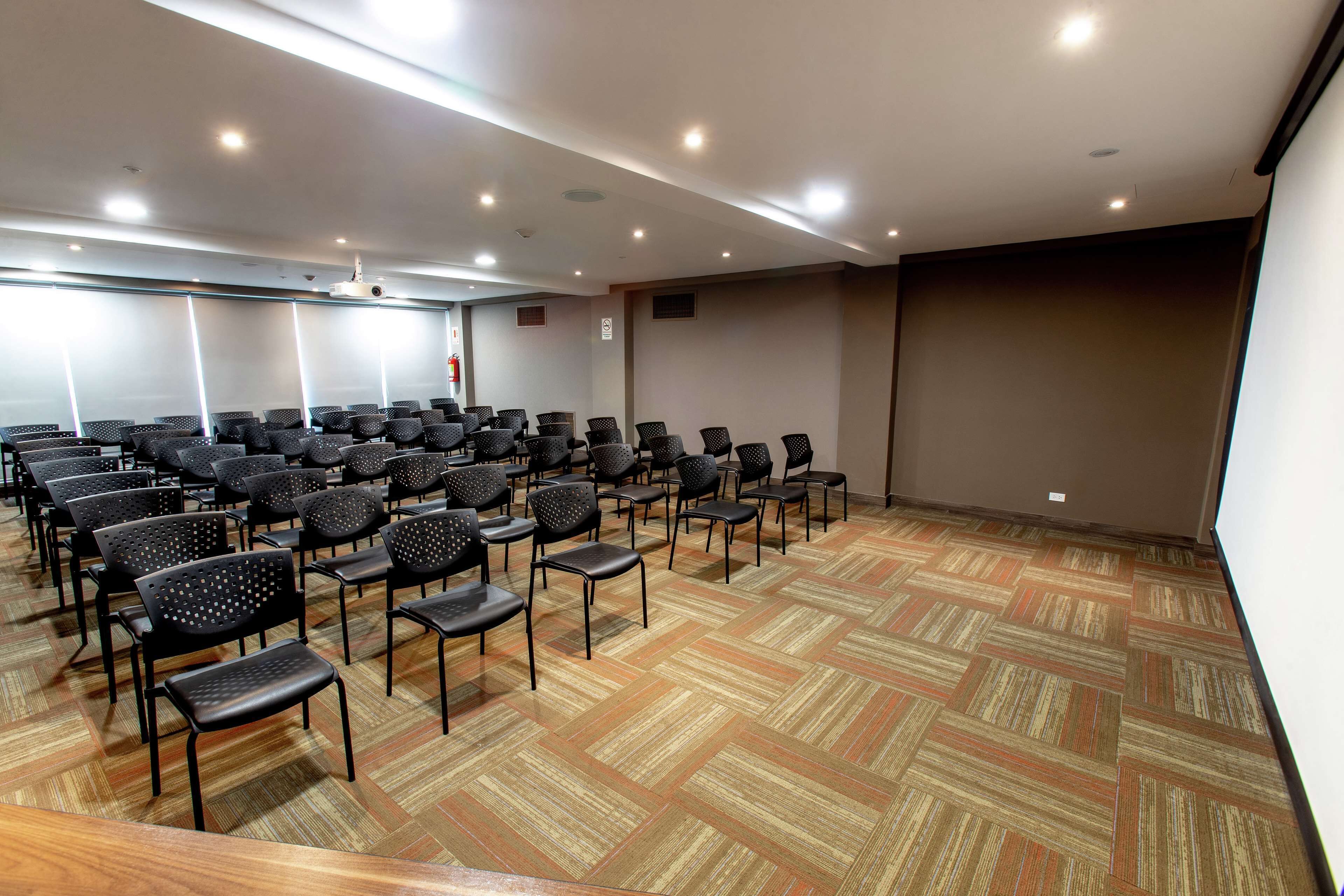 Une salle de conférence avec des rangées de chaises, un sol recouvert de moquette et un écran de projection.