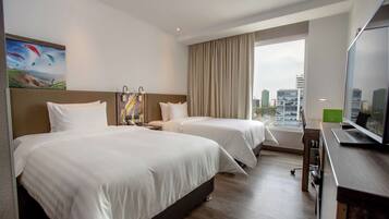 Habitación de hotel con dos camas, un escritorio con computadora, un ventanal con vista a la ciudad y un cuadro en la pared.