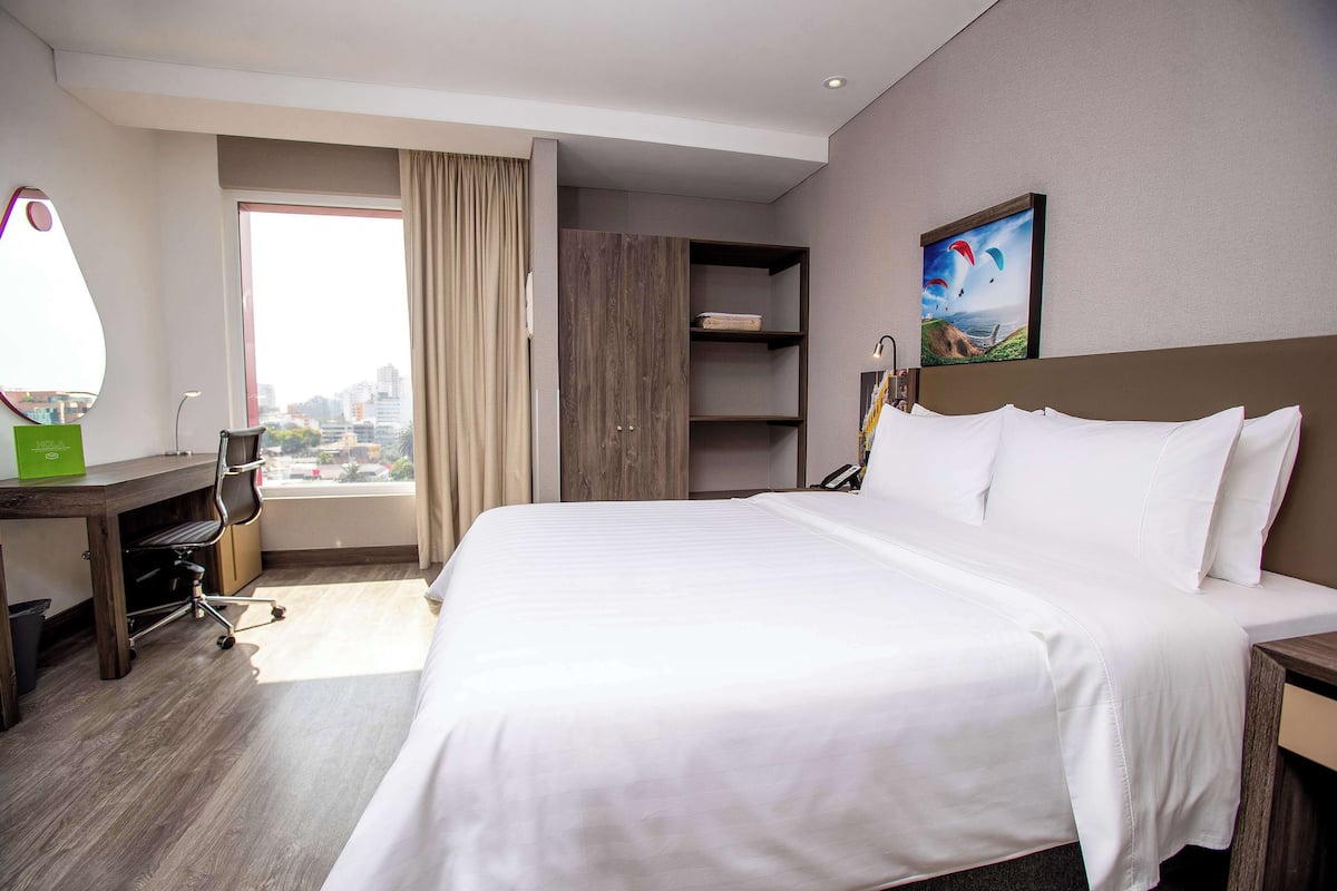 A hotel room with a large bed, a desk with a chair, a wardrobe, and a window with a city view.