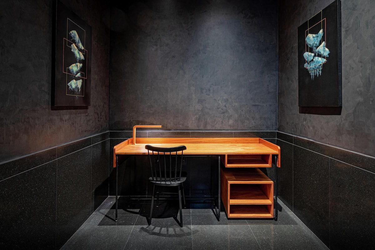 A modern, dark-themed room with a wooden desk and a black chair. There are two framed artworks on the walls.