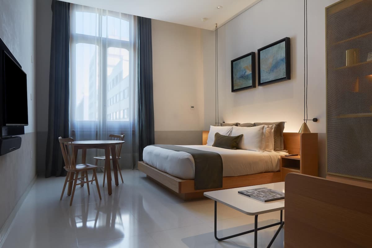 A modern hotel room with a bed, bedside table, and a dining area with chairs.