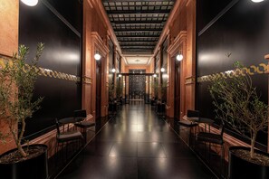 A corridor with a polished floor, flanked by potted plants and chairs, leading to a central area with a decorative ceiling and a door at the end. - Umbral, a Curio Collection by Hilton - Historical Hotel (Mexico City)