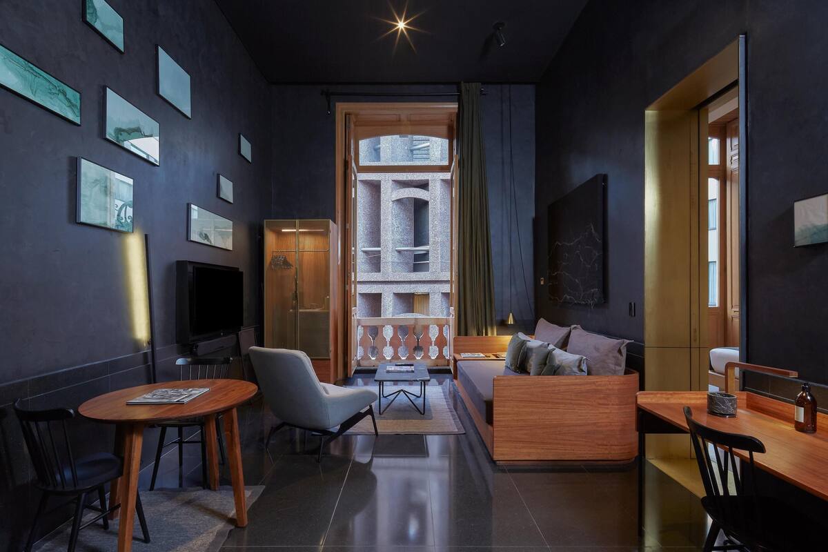 A modern living room with a sofa, coffee table, and chairs. There is a TV and a wooden cabinet. The room has a large window with curtains.