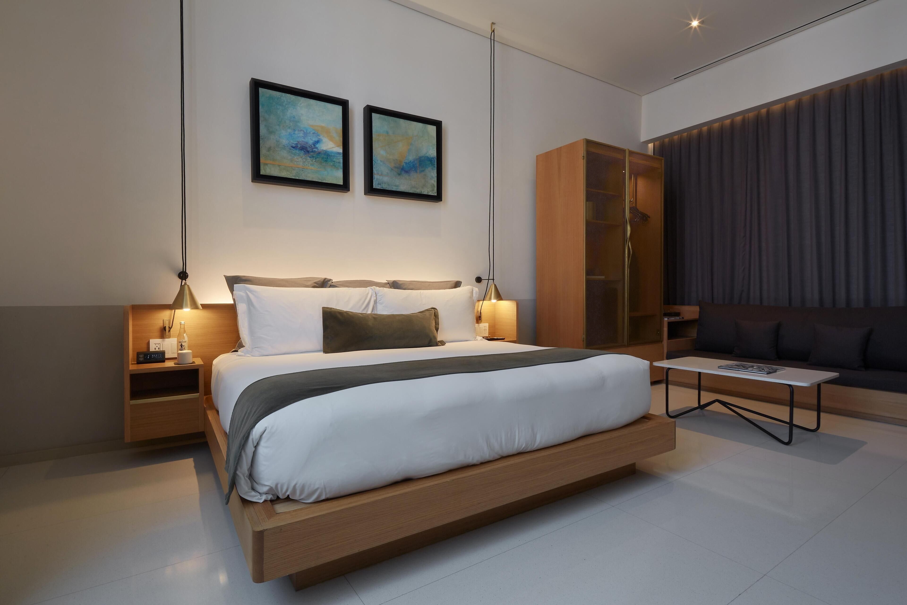 A modern bedroom with a large bed, bedside tables, a nightstand, a wooden wardrobe, a sofa, and two abstract paintings on the wall.