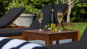 Een houten tafel met twee champagneglazen en een fles, omringd door loungestoelen en groen.
