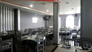 A spacious dining area with multiple tables and chairs, a television mounted on the wall, and large windows allowing natural light. - Hotel Restaurante América (A Estrada)