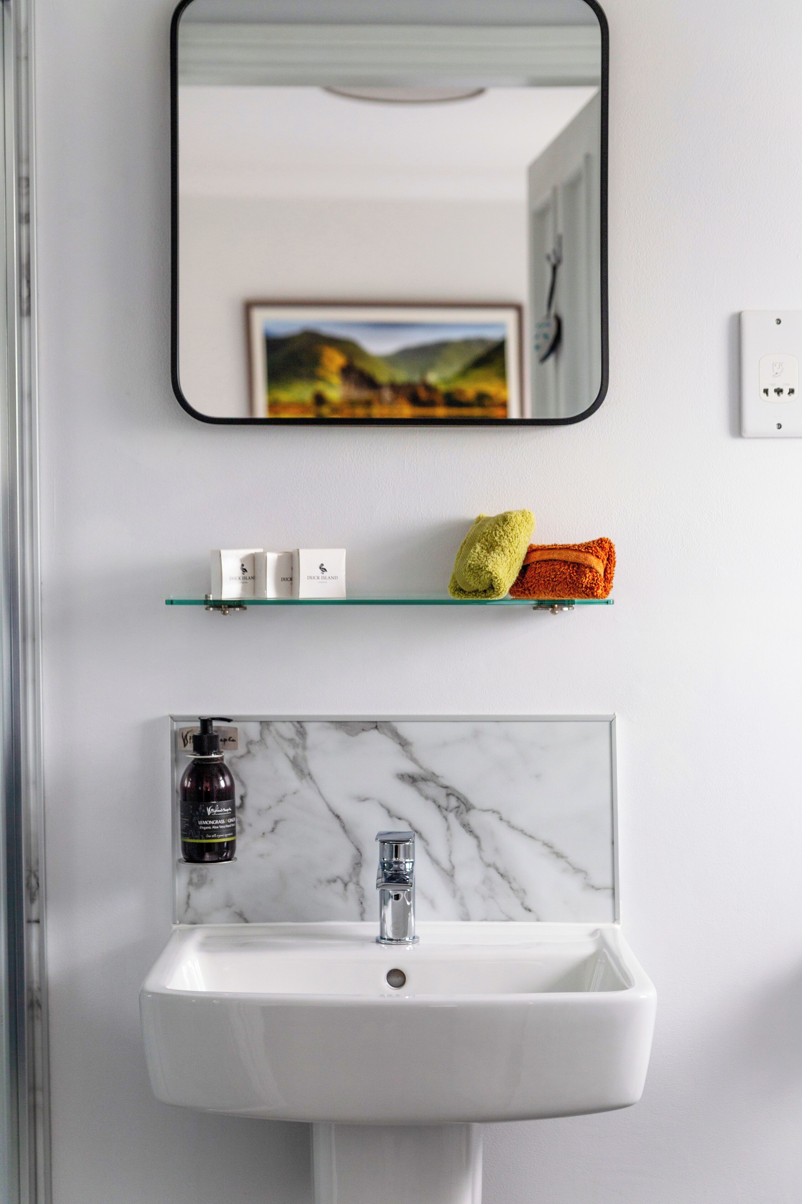 Ein Badezimmer mit einem weißen Waschbecken, einer Glasablage für Pflegeprodukte und einem gerahmten Bild an der Wand.