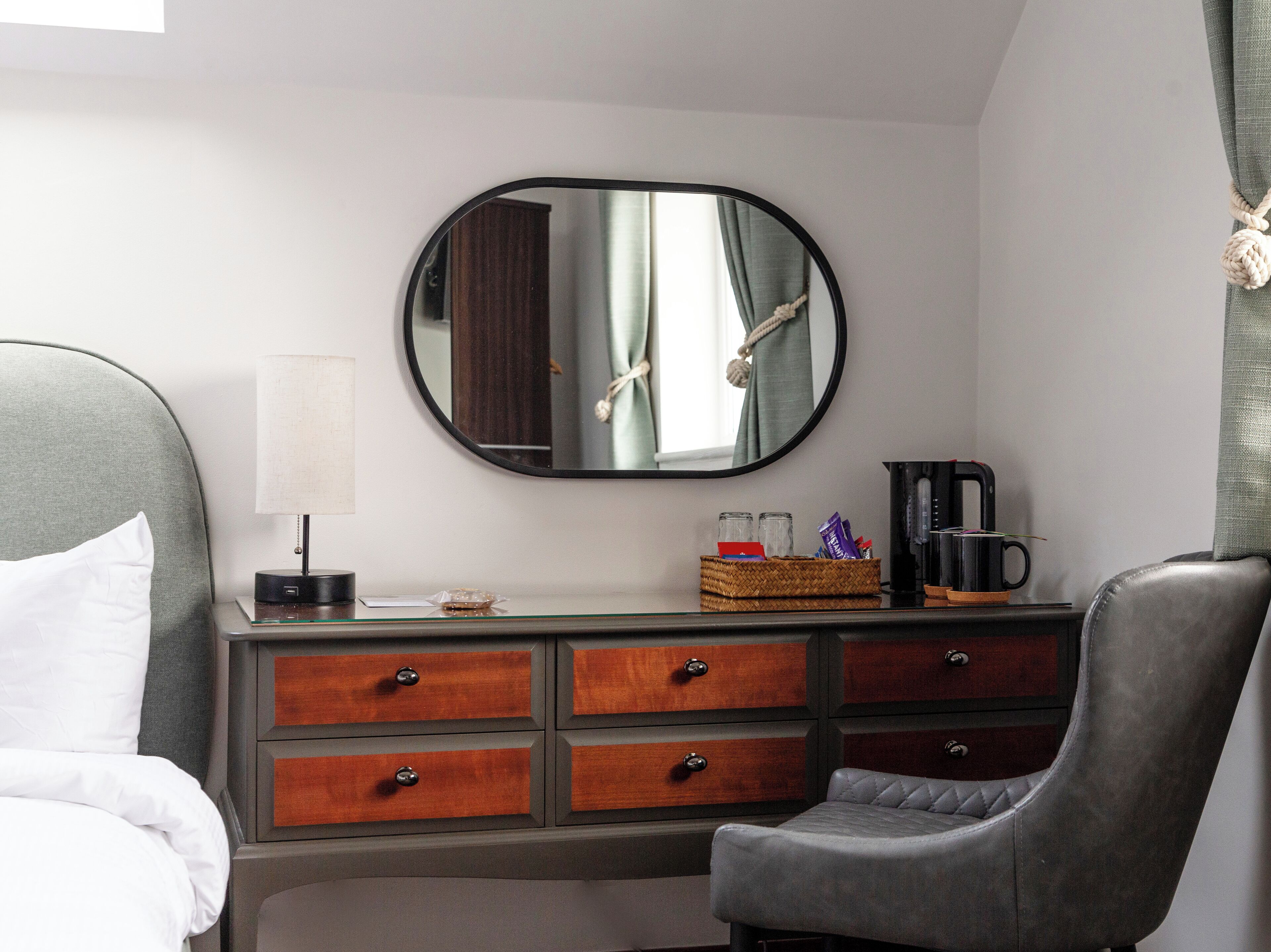 A hotel room with a bed, a desk with drawers, a chair, a coffee maker, a lamp, and a mirror on the wall.