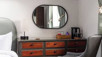 A hotel room with a bed, a desk with drawers, a chair, a coffee maker, a lamp, and a mirror on the wall.