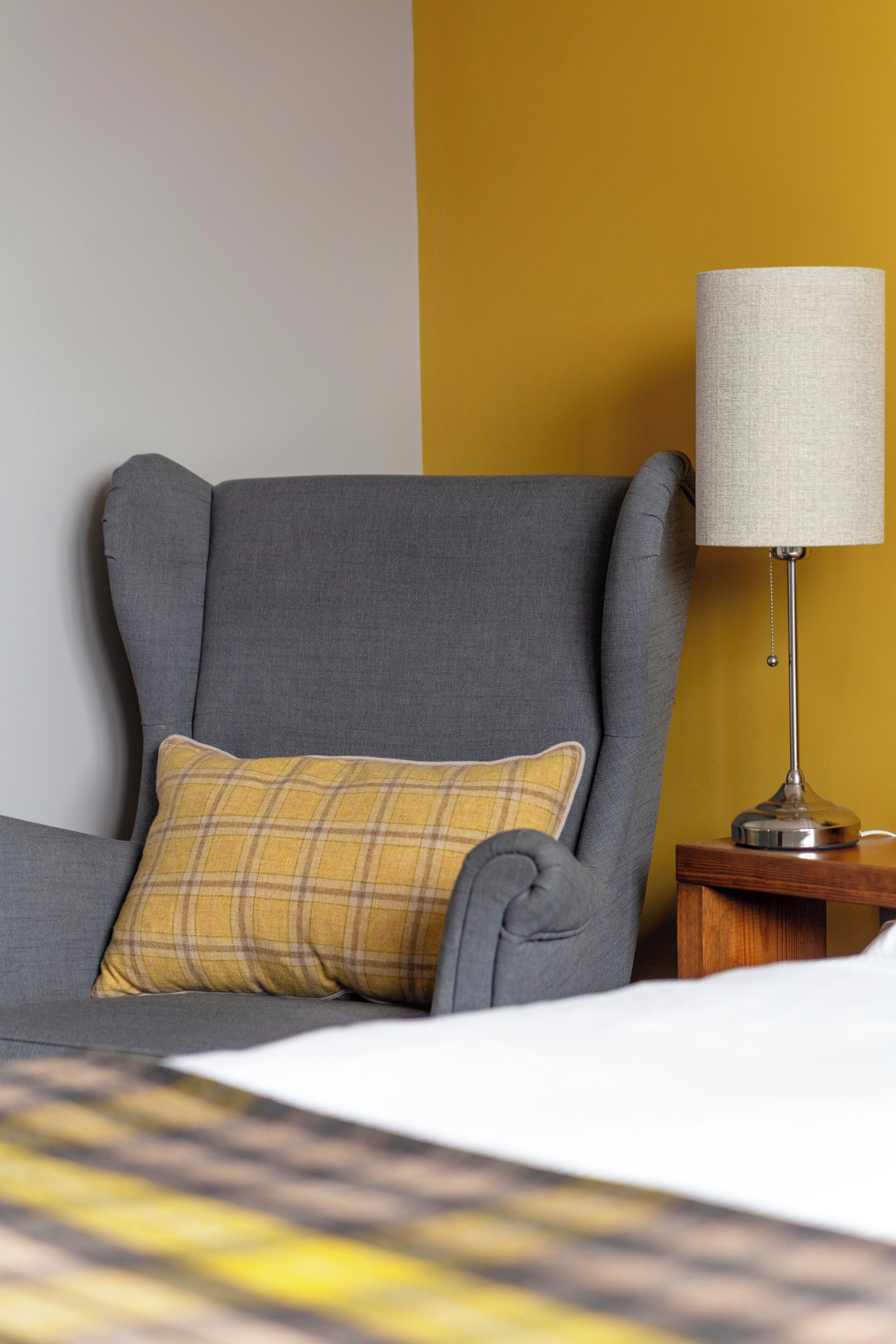 Un fauteuil gris avec un coussin à carreaux, une lampe sur une table de chevet en bois et un lit recouvert d’une couvre-lit à motifs blancs et jaunes.