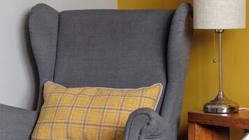 Un fauteuil gris avec un coussin à carreaux, une lampe sur une table de chevet en bois et un lit recouvert d’une couvre-lit à motifs blancs et jaunes.