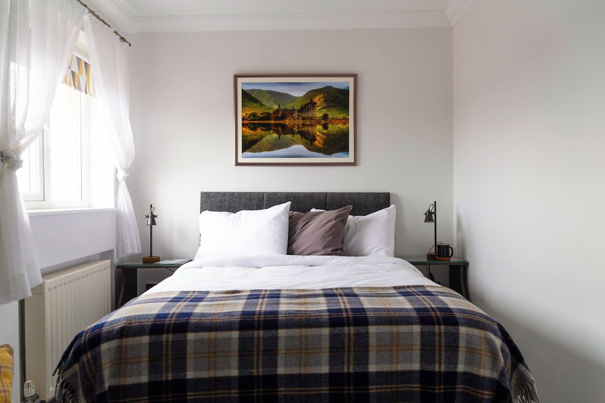 A neatly made bed with a plaid blanket, a framed landscape painting above it, and a bedside table with a lamp and a mug.