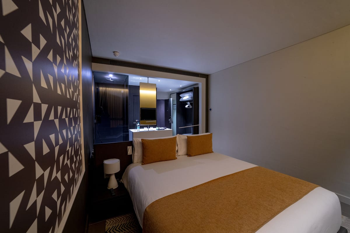 Habitación de hotel con una cama grande, una mesita de noche con lámpara, un espejo y una pared con diseño geométrico.