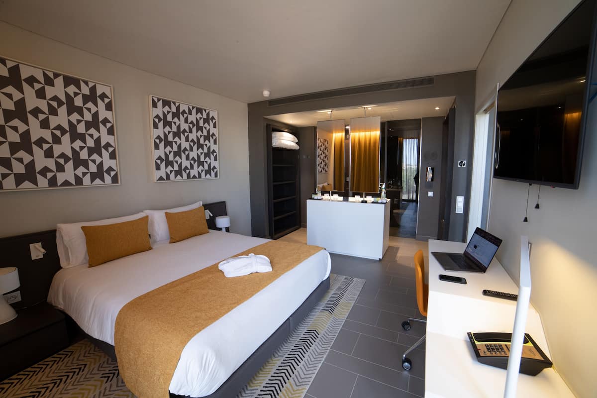 Una habitación de hotel moderna con una cama grande, un escritorio con una computadora portátil, un televisor y un baño visible a través de una puerta abierta.