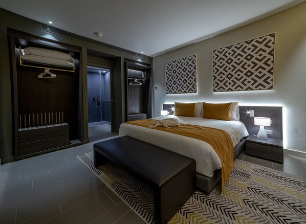 A modern hotel room with a large bed, a bench, a walk-in closet, and a shower area.