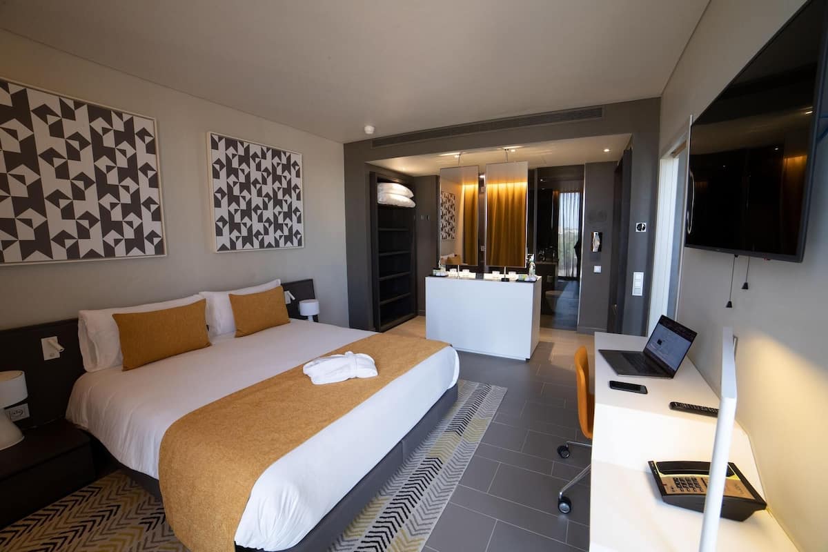 Una habitación de hotel moderna con una cama grande, un escritorio con una computadora portátil, un televisor y un baño con espejo y lavamanos.