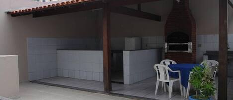 An outdoor patio with a brick oven, white chairs, and a blue tablecloth.