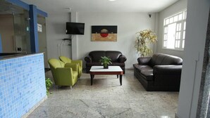 Un vestíbulo con suelo de baldosas, un mostrador de recepción, dos sillones verdes, un sofá de cuero marrón, una mesa de centro de madera, una planta en maceta y un televisor de pantalla plana montado en la pared.