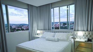 A hotel room with a large bed, a nightstand with a lamp, and a view of a cityscape through large windows. - Paracatu Plaza Hotel (Paracatu)