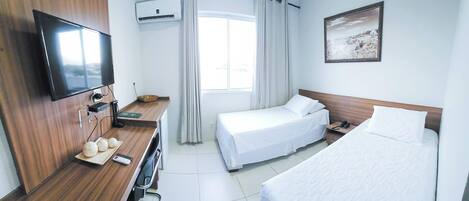 A hotel room with a wooden desk, a flat-screen TV, two beds, a window with curtains, and a painting on the wall.