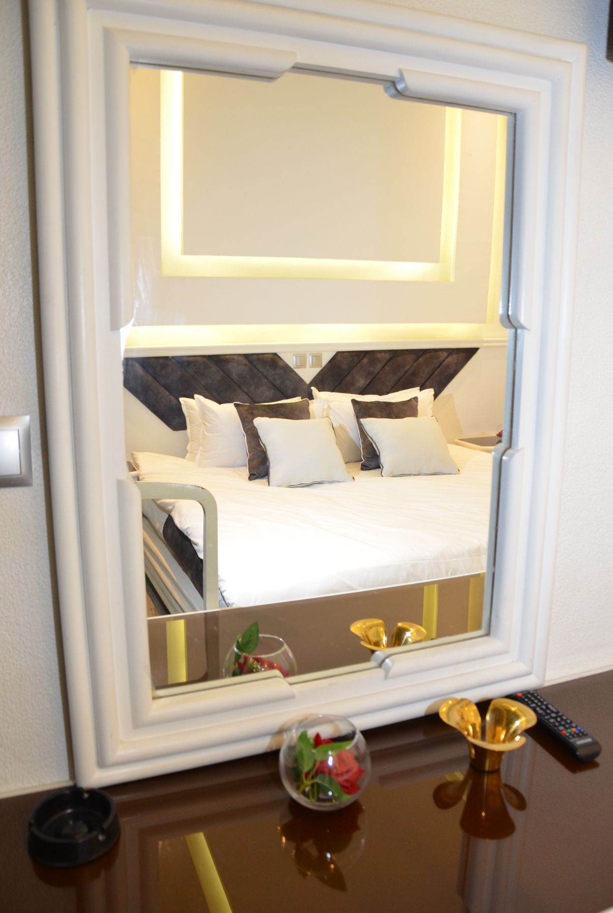 A hotel room with a bed, a bedside table with a remote control, and a mirror reflecting the room.