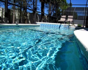 Une piscine aux eaux claires, bordée de carreaux, entourée d’arbres et avec un bâtiment visible au loin.